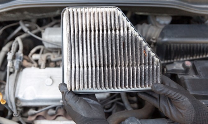 mechanic-holding-dirty-air-filter Someone wearing rubber gloves holding up a dirty air filter above an open engine area of a vehicle in the background.